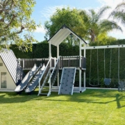 A white A frame playhouse with slides and swings from King Swings in a backyard with lots of trees and shade