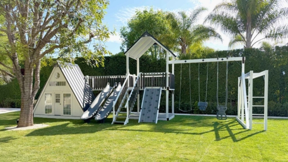 A white A frame playhouse with slides and swings from King Swings in a backyard with lots of trees and shade